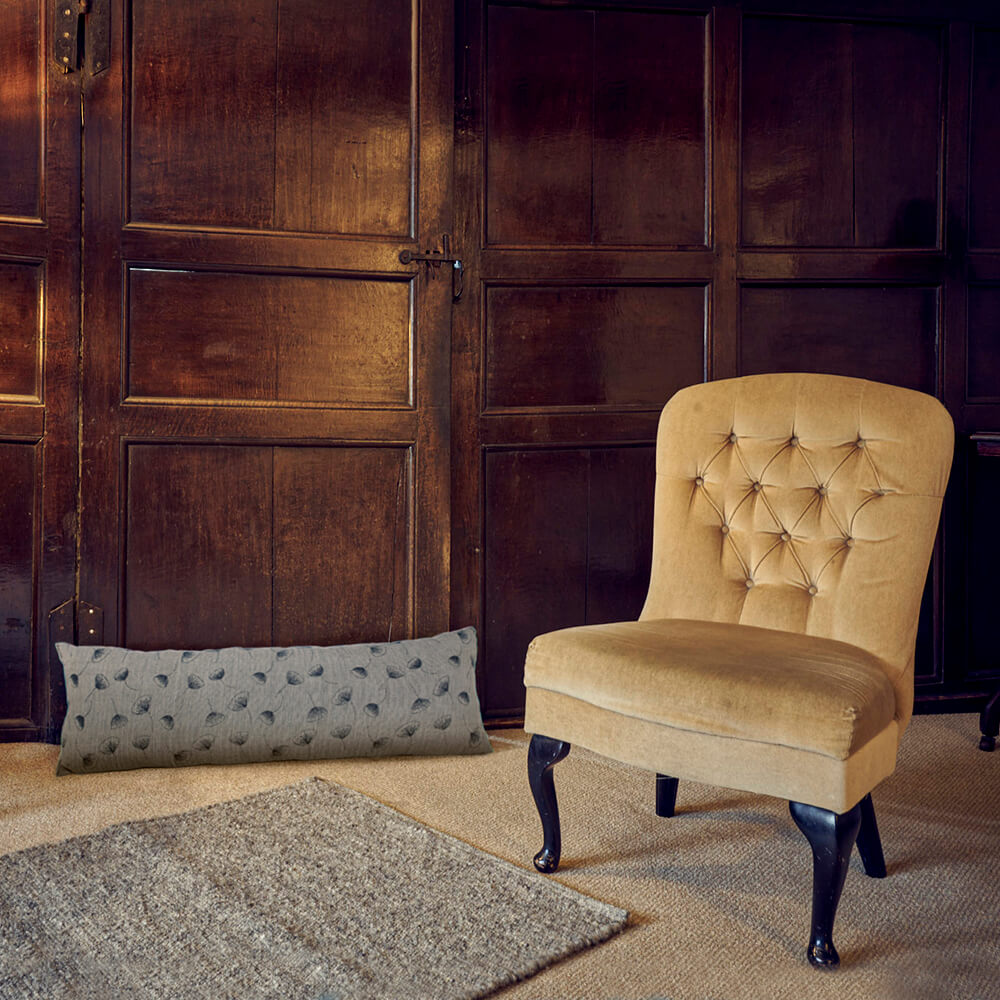 A classic tufted armchair upholstered in a warm beige velvet fabric, set against a dark wood paneled wall. A matching textured throw pillow and a gray area rug are placed on the floor in front of the chair.