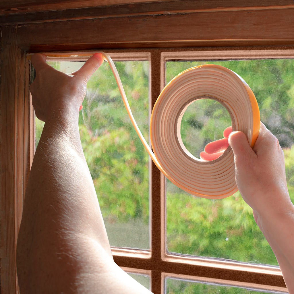 A person's hands apply draught seal tape to a wooden window frame, with a roll of the seal held up against a window and green foliage visible outside.
