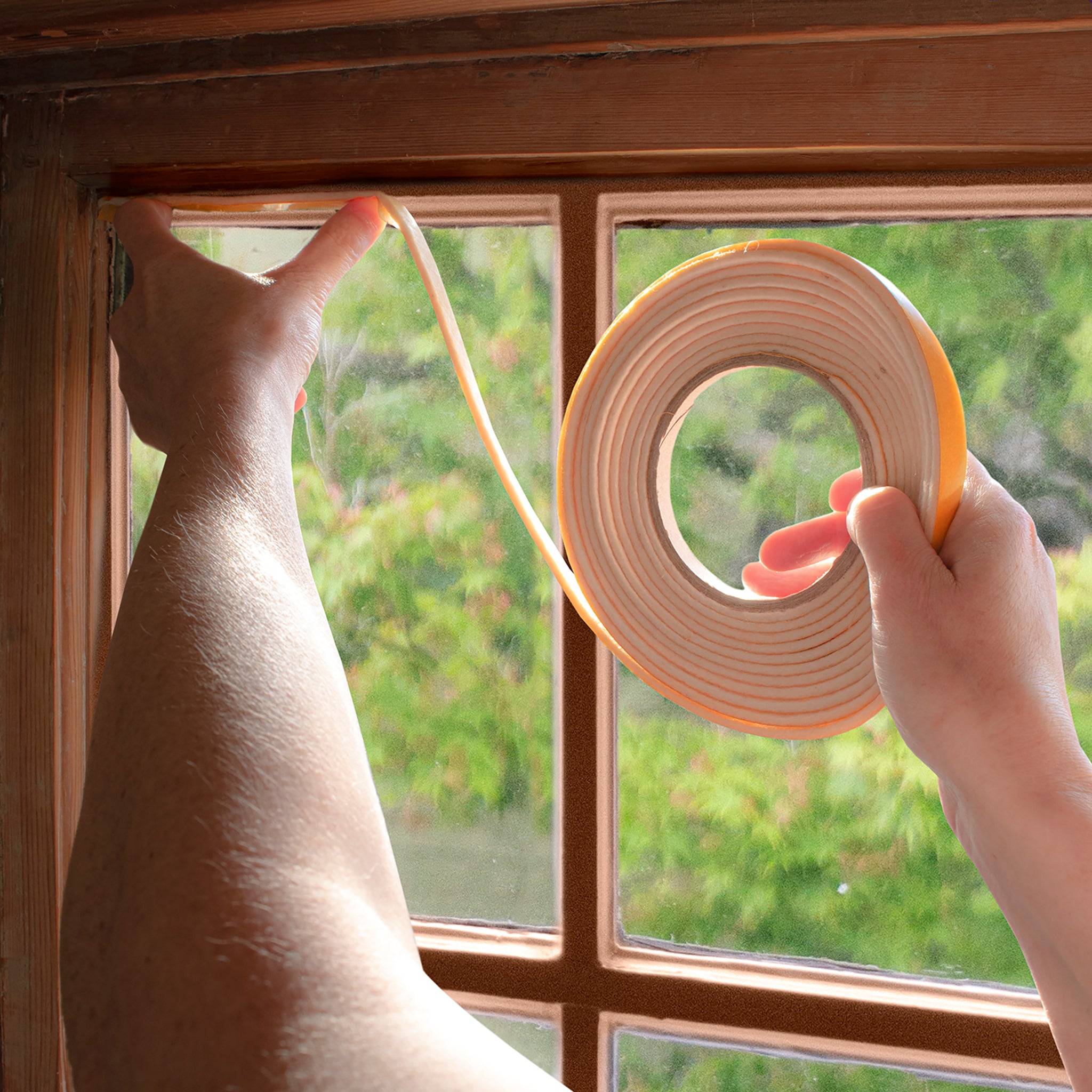 A person's hands apply draught seal tape to a wooden window frame, with a roll of the seal held up against a window and green foliage visible outside.
