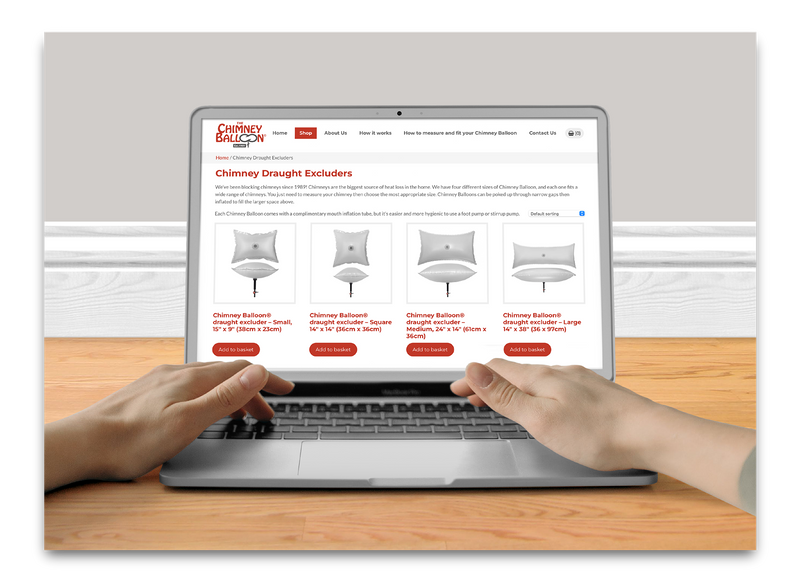 A person's hands typing on a silver laptop computer on a wooden desk, with the laptop screen displaying a website for Chimney Draft Excluders.