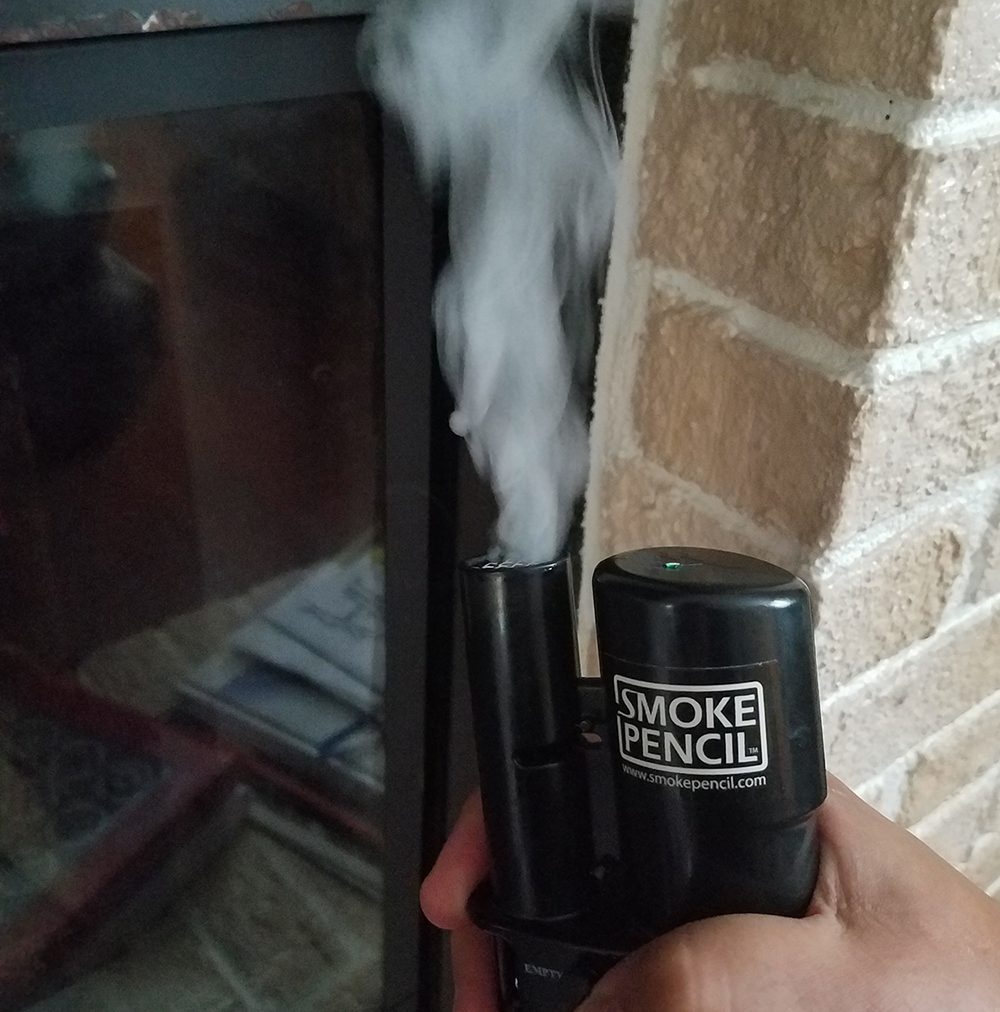 A hand holding a black smoke pencil that is emitting a large cloud of white smoke next to a brick fireplace.