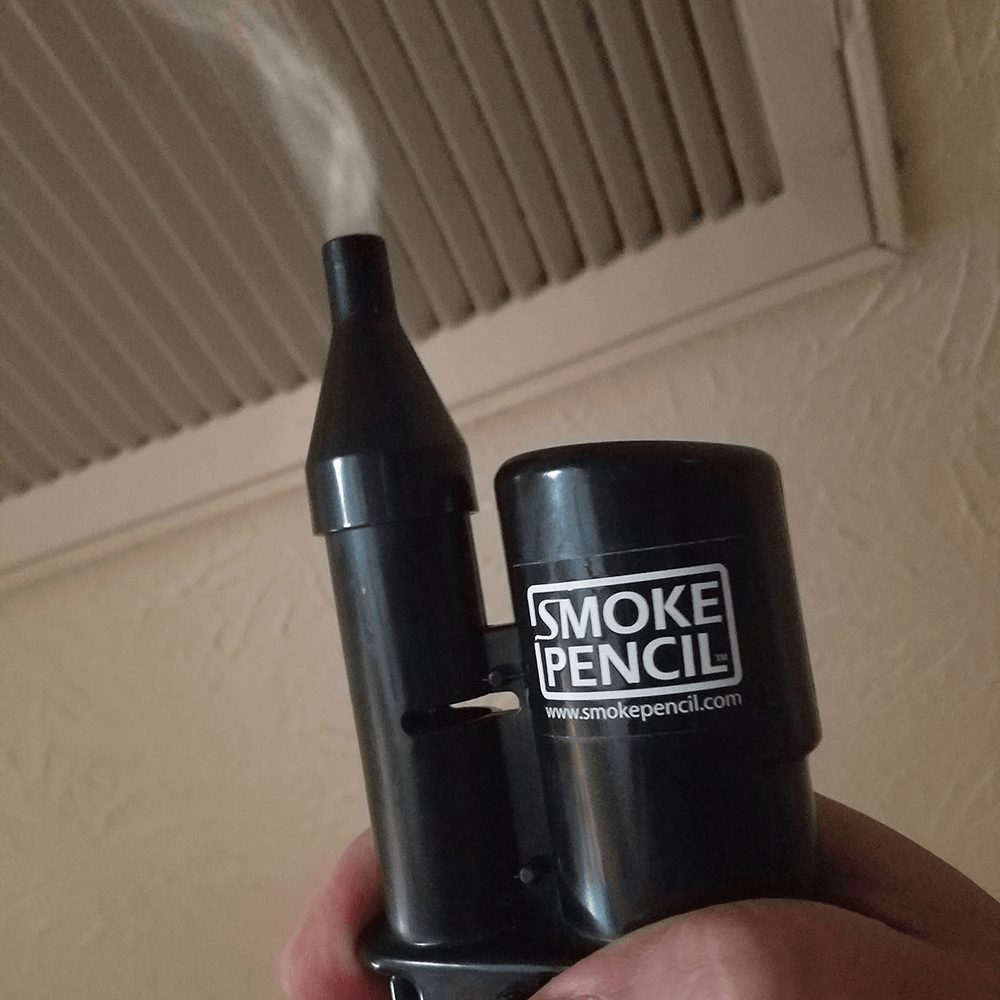 A black Smoke Pencil electronic smoking device with a visible misting effect from the top nozzle, held in a hand against a beige wall and vent background.