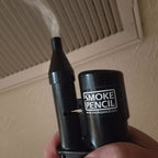 A black Smoke Pencil electronic smoking device with a visible misting effect from the top nozzle, held in a hand against a beige wall and vent background.
