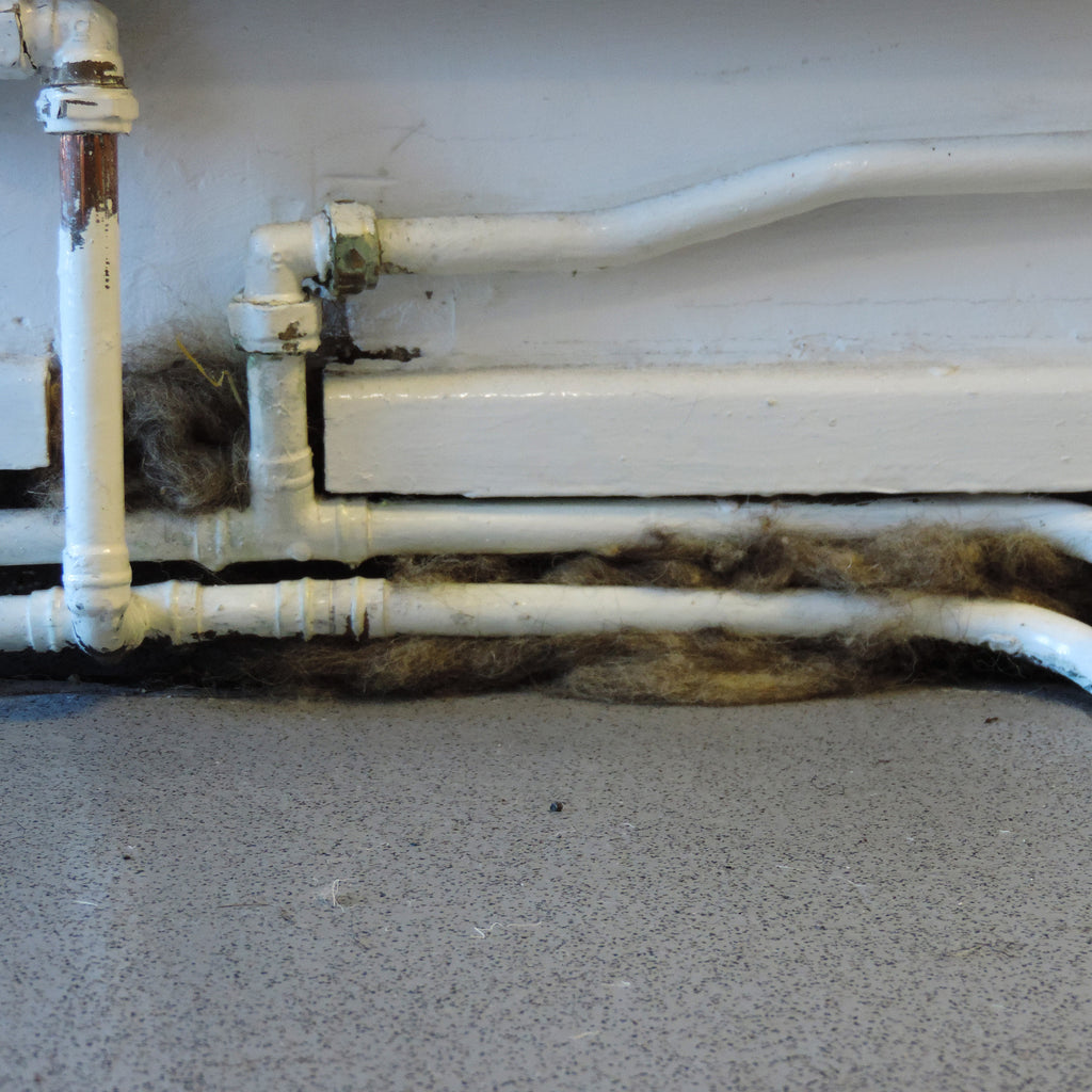 White PVC plumbing pipes installed along the base of a white wall above a grey speckled floor, with a thick layer of dark brown and grey wool insulation pushed between the pipes.